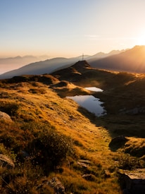 A serene landscape with rolling hills covered in grass, gently illuminated by the golden light of a sunset. A small body of water reflects the sky, and a cross stands on top of a hill, suggesting a tranquil, contemplative setting. Mountain peaks are visible in the background, shrouded in a light mist, adding depth to the scene.