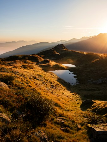 A serene landscape with rolling hills covered in grass, gently illuminated by the golden light of a sunset. A small body of water reflects the sky, and a cross stands on top of a hill, suggesting a tranquil, contemplative setting. Mountain peaks are visible in the background, shrouded in a light mist, adding depth to the scene.