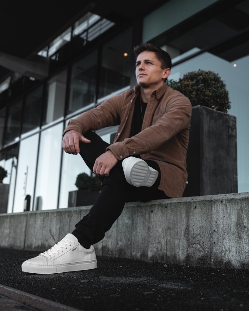 man in brown leather jacket and white nike sneakers sitting on concrete wall during daytime