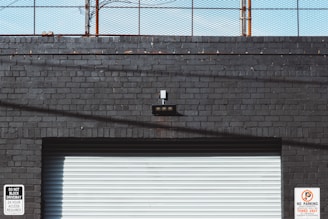 A gray brick wall with a rolling shutter door occupies the lower part of the image. Two signs are present: one on the left reads 'Do Not Block Driveway 24 Hour Access Required,' and another on the right warns that unauthorized vehicles will be towed at the owner's expense. Above the door is a small black light fixture with five bulbs. A rusty fence runs along the top of the wall against a blue sky.