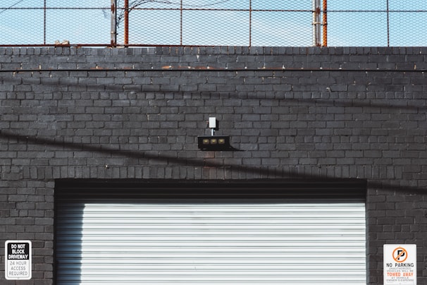 A gray brick wall with a rolling shutter door occupies the lower part of the image. Two signs are present: one on the left reads 'Do Not Block Driveway 24 Hour Access Required,' and another on the right warns that unauthorized vehicles will be towed at the owner's expense. Above the door is a small black light fixture with five bulbs. A rusty fence runs along the top of the wall against a blue sky.