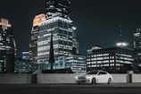 A white car is parked on a rooftop with a backdrop of tall, illuminated skyscrapers in a city at night. The buildings are brightly lit with various patterns of windows, creating a vibrant urban atmosphere.