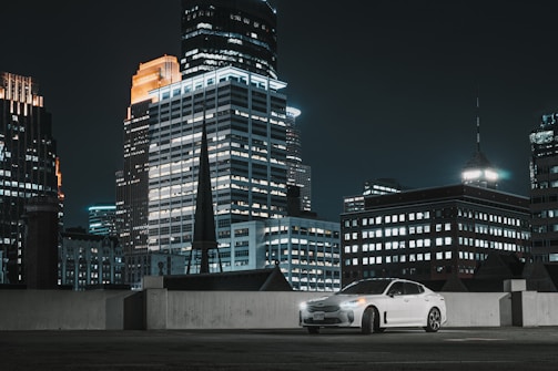 A white car is parked on a rooftop with a backdrop of tall, illuminated skyscrapers in a city at night. The buildings are brightly lit with various patterns of windows, creating a vibrant urban atmosphere.