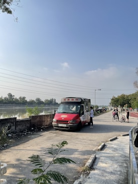 An ice cream truck is parked on a pathway beside a body of water. A person stands near the truck, appearing to make a purchase. In the background, there are more people and bicycles. The sky is clear and blue, contributing to a sunny day ambiance. Trees and greenery line the area on the right.