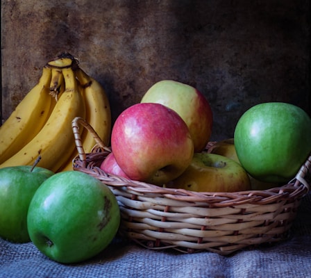 A woven basket filled with a variety of apples sits on a textured fabric surface. Beside the basket, a bunch of ripe bananas is positioned upright. The apples display shades of red, yellow, and green, adding a colorful contrast to the darker, rustic background.