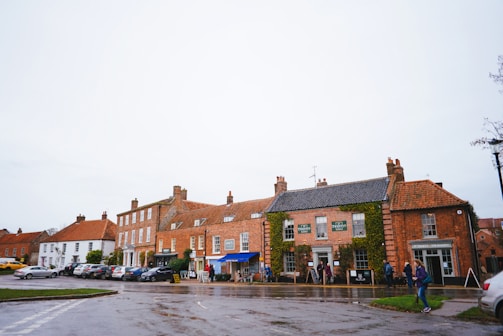 A quaint village street featuring a row of brick and whitewashed buildings with red-tiled roofs. Several signs and a blue awning suggest small shops and possibly a pub. A number of parked cars line the street, and a few people are walking along the sidewalk. The ground appears wet, indicating recent rain, under a pale gray sky.