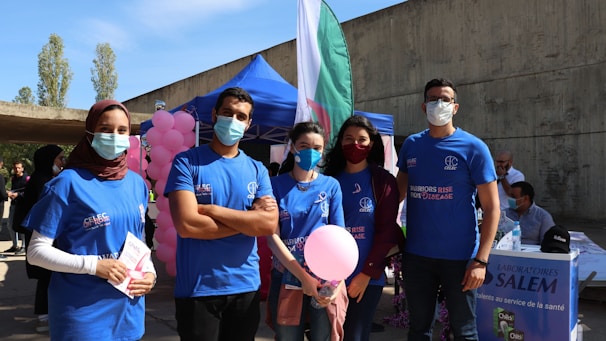 A group of five people, wearing blue shirts and medical masks, stand outdoors at a health event. Some are holding informational pamphlets and a pink balloon, with a background that includes a booth displaying a sign for 'Laboratoires Salem' and pink balloons. The setting appears to be a health awareness or fundraising event. A large flag and tents are visible behind them.
