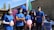 A group of five people, wearing blue shirts and medical masks, stand outdoors at a health event. Some are holding informational pamphlets and a pink balloon, with a background that includes a booth displaying a sign for 'Laboratoires Salem' and pink balloons. The setting appears to be a health awareness or fundraising event. A large flag and tents are visible behind them.