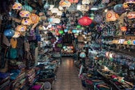 A vibrant and colorful shop filled with a variety of intricately designed lamps hanging from the ceiling. Shelves on both sides are packed with an array of goods, including bags, textiles, and decorative items. The mosaic patterns on the lamps create an eclectic and exotic atmosphere.