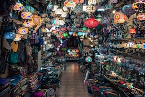 A vibrant and colorful shop filled with a variety of intricately designed lamps hanging from the ceiling. Shelves on both sides are packed with an array of goods, including bags, textiles, and decorative items. The mosaic patterns on the lamps create an eclectic and exotic atmosphere.