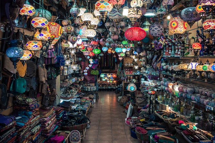 A vibrant and colorful shop filled with a variety of intricately designed lamps hanging from the ceiling. Shelves on both sides are packed with an array of goods, including bags, textiles, and decorative items. The mosaic patterns on the lamps create an eclectic and exotic atmosphere.