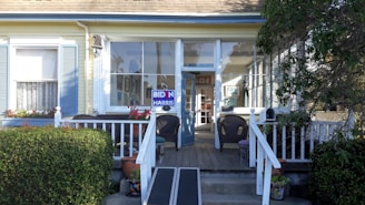 A residential front porch features a few wicker chairs and a wooden ramp leading up to a glass entrance door. The exterior of the house is painted in pale yellow with light blue shutters and white trim. There is a political sign reading 'BIDEN HARRIS' placed prominently on the porch rail. Potted plants and flowers add some color to the scene. Large windows behind the seating offer a glimpse into the interior.