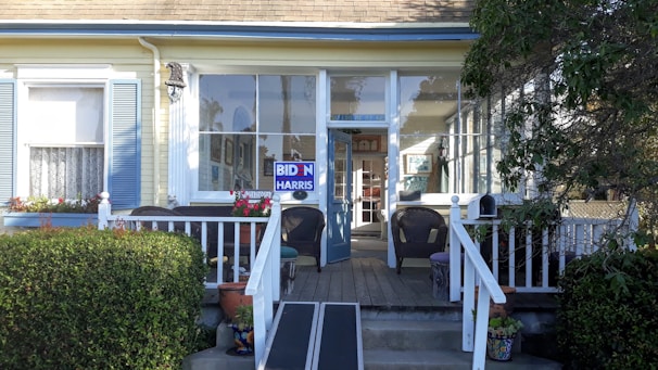 A residential front porch features a few wicker chairs and a wooden ramp leading up to a glass entrance door. The exterior of the house is painted in pale yellow with light blue shutters and white trim. There is a political sign reading 'BIDEN HARRIS' placed prominently on the porch rail. Potted plants and flowers add some color to the scene. Large windows behind the seating offer a glimpse into the interior.
