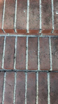 A close-up view of a brick pavement with multiple rectangular red bricks aligned in rows. The gaps between the bricks are filled with a light gray mortar or grout.