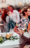 A hand with polished nails holding a small skewer appetizer with basil, cheese, and a cherry tomato in a social gathering setting. In the background, a group of people engages in conversation, with blurred figures and bokeh lighting. The scene is vibrant and lively, featuring a white table adorned with leaves, citrus fruits, and partially filled wine glasses.
