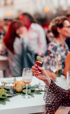 A hand with polished nails holding a small skewer appetizer with basil, cheese, and a cherry tomato in a social gathering setting. In the background, a group of people engages in conversation, with blurred figures and bokeh lighting. The scene is vibrant and lively, featuring a white table adorned with leaves, citrus fruits, and partially filled wine glasses.