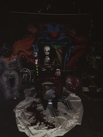The image depicts a dark, eerie setting with an ominous clown theme. In the center, there is a figure dressed in a sinister clown costume, sitting in front of a backdrop featuring a menacing clown face with sharp teeth and vivid colors. Surrounding the central figure are additional unsettling elements, such as props and decorations that enhance the horror theme, including fake blood and crime scene tape. The lighting is dim, contributing to a spooky atmosphere.