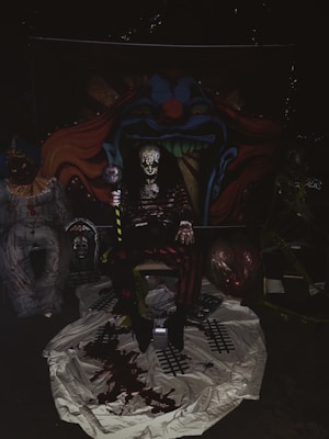 The image depicts a dark, eerie setting with an ominous clown theme. In the center, there is a figure dressed in a sinister clown costume, sitting in front of a backdrop featuring a menacing clown face with sharp teeth and vivid colors. Surrounding the central figure are additional unsettling elements, such as props and decorations that enhance the horror theme, including fake blood and crime scene tape. The lighting is dim, contributing to a spooky atmosphere.