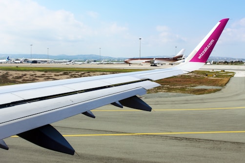 An airplane wing is visible in the foreground with a purple winglet marked 'wizzair.com'. The scene captures a runway with multiple aircraft parked in the distance. The sky is partly cloudy, and there are patches of greenery alongside the runway.