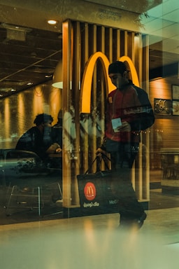 A person wearing a cap and carrying a branded bag is leaving a fast-food restaurant. The iconic logo is prominently displayed in the background, indicating the location. Inside, another person is seated, creating a casual and everyday setting.