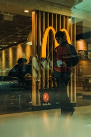 A person wearing a cap and carrying a branded bag is leaving a fast-food restaurant. The iconic logo is prominently displayed in the background, indicating the location. Inside, another person is seated, creating a casual and everyday setting.