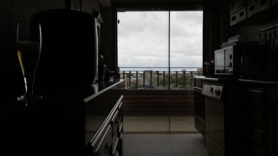 A modern kitchen with dark tones, featuring a countertop with kitchen appliances and utensils. The room is dimly lit, with a large window in the background overlooking a cityscape under a cloudy sky. The silhouette of a glass with a drink is visible on the counter.