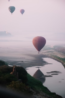 Hot air balloons float gently over a serene landscape with a river reflecting the silhouette of one of the balloons. The scene is enveloped in a mist, giving it a dreamlike quality. A distant view of a pagoda can be seen on the horizon, adding to the tranquil and exotic atmosphere.