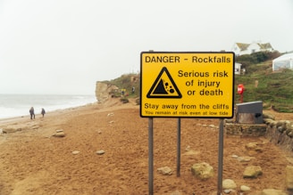A coastal landscape featuring a large yellow warning sign in the foreground. The sign alerts about the dangers of rockfalls and warns of the serious risk of injury or death, advising people to stay away from the cliffs. The background includes a sandy beach with scattered rocks, a few people walking near the shoreline, and cliffs alongside the sea.