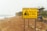 A coastal landscape featuring a large yellow warning sign in the foreground. The sign alerts about the dangers of rockfalls and warns of the serious risk of injury or death, advising people to stay away from the cliffs. The background includes a sandy beach with scattered rocks, a few people walking near the shoreline, and cliffs alongside the sea.
