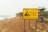 A coastal landscape featuring a large yellow warning sign in the foreground. The sign alerts about the dangers of rockfalls and warns of the serious risk of injury or death, advising people to stay away from the cliffs. The background includes a sandy beach with scattered rocks, a few people walking near the shoreline, and cliffs alongside the sea.