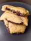 A stack of homemade cookies filled with melted chocolate is placed on a dark plate. The cookies have a golden-brown crust, and the melted chocolate is visibly oozing out from the center, creating a tempting and delicious appearance.