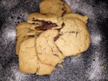 A stack of freshly baked cookies is arranged on a dark surface dusted with flour. The cookies are golden brown with visible chunks of melted chocolate.
