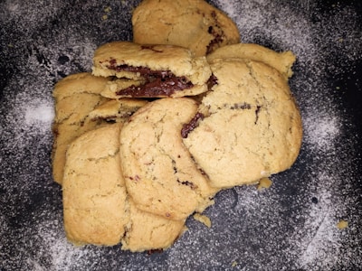 A stack of freshly baked cookies is arranged on a dark surface dusted with flour. The cookies are golden brown with visible chunks of melted chocolate.