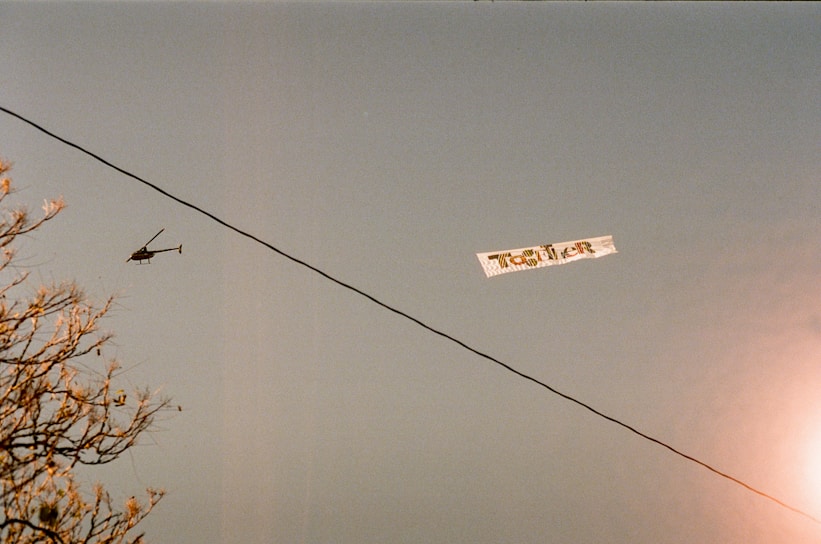 A helicopter flies through the sky towing an advertising banner. Near the bottom left, tree branches with autumn-colored leaves fill part of the frame. A black utility wire crosses the scene diagonally, creating a sense of division in the sky.