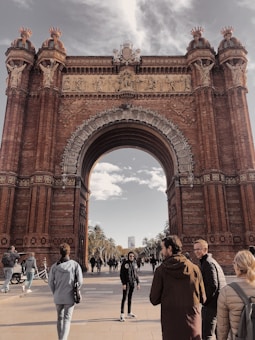 A large, ornate archway constructed from reddish-brown bricks with intricate carvings and sculptures stands prominently. The structure features classical design elements and decorative reliefs. People are walking and standing around the archway; some are conversing, creating a lively atmosphere. The sky is partly cloudy, and palm trees line the pathway behind the arch.