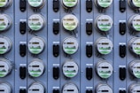 A grid of multiple electric utility meters mounted on a panel. The arrangement is orderly, with each meter featuring a transparent cover and a digital display, some of which have protective black covers next to them.