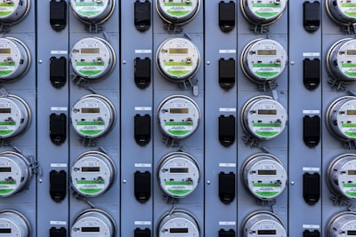 A grid of multiple electric utility meters mounted on a panel. The arrangement is orderly, with each meter featuring a transparent cover and a digital display, some of which have protective black covers next to them.