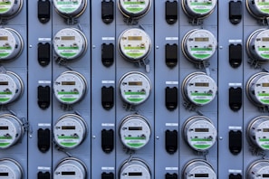 A grid of multiple electric utility meters mounted on a panel. The arrangement is orderly, with each meter featuring a transparent cover and a digital display, some of which have protective black covers next to them.