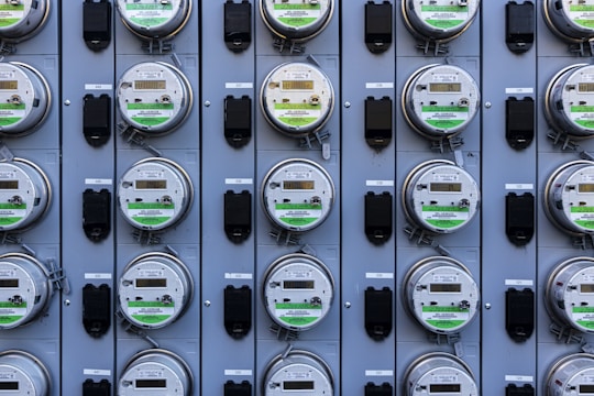 A grid of multiple electric utility meters mounted on a panel. The arrangement is orderly, with each meter featuring a transparent cover and a digital display, some of which have protective black covers next to them.