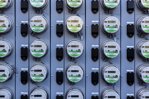 A grid of multiple electric utility meters mounted on a panel. The arrangement is orderly, with each meter featuring a transparent cover and a digital display, some of which have protective black covers next to them.