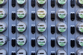 A grid of multiple electric utility meters mounted on a panel. The arrangement is orderly, with each meter featuring a transparent cover and a digital display, some of which have protective black covers next to them.