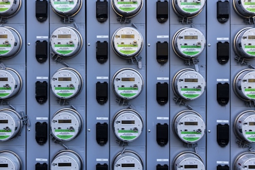A grid of multiple electric utility meters mounted on a panel. The arrangement is orderly, with each meter featuring a transparent cover and a digital display, some of which have protective black covers next to them.