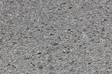 A close-up view of a rough asphalt surface composed of small, uneven gravel and stone pieces. The texture is coarse and heterogeneous, with various shades of gray and occasional hints of brown.