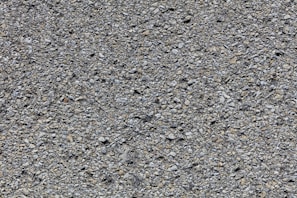 A close-up view of a rough asphalt surface composed of small, uneven gravel and stone pieces. The texture is coarse and heterogeneous, with various shades of gray and occasional hints of brown.
