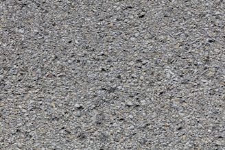 A close-up view of a rough asphalt surface composed of small, uneven gravel and stone pieces. The texture is coarse and heterogeneous, with various shades of gray and occasional hints of brown.