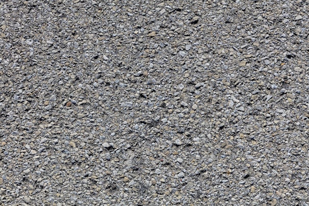 A close-up view of a rough asphalt surface composed of small, uneven gravel and stone pieces. The texture is coarse and heterogeneous, with various shades of gray and occasional hints of brown.