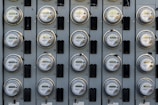 A grid of electric meters is mounted on a grey panel, arranged in neat, aligned rows and columns. Each meter has a circular, glass-covered display with numerical readouts and labels, encased in a metallic frame.