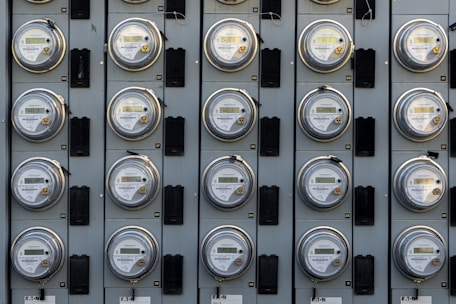 A grid of electric meters is mounted on a grey panel, arranged in neat, aligned rows and columns. Each meter has a circular, glass-covered display with numerical readouts and labels, encased in a metallic frame.