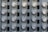 A grid of electric meters is mounted on a grey panel, arranged in neat, aligned rows and columns. Each meter has a circular, glass-covered display with numerical readouts and labels, encased in a metallic frame.