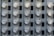 A grid of electric meters is mounted on a grey panel, arranged in neat, aligned rows and columns. Each meter has a circular, glass-covered display with numerical readouts and labels, encased in a metallic frame.